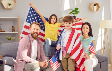 Portrait of a young happy American family with two kids sitting on sofa at home with flag of united...