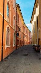 Narrow alley between historic buildings with arched windows and orange walls on a sunny day. Trash bins lined up on the side, blue sky in the background.