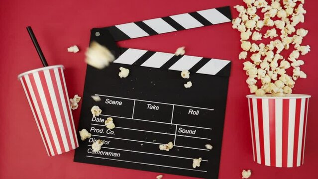 Popcorn popcorn falls on Cinema clapper board with soda cup, video production, film theater treat, movie tickets, cinematography entertainment, showtime snack, red background