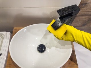 close-up female hand in yellow gloves cleaning faucet of bathroom sink with sponge