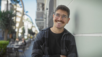 A smiling young hispanic man with a moustache standing in a sunlit urban street