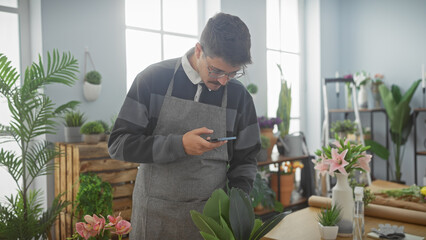 Hispanic man with glasses and apron using smartphone in a flower shop surrounded by plants and...