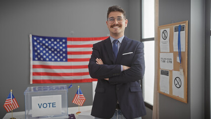 A confident young hispanic man stands arms crossed in a voting center with a us flag backdrop