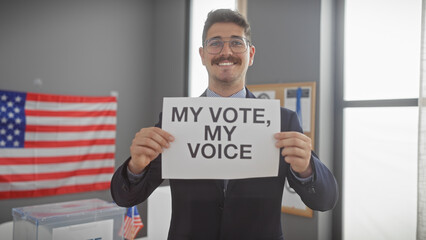 Smiling young hispanic man holding 'my vote, my voice' sign in a voting center with us flag.