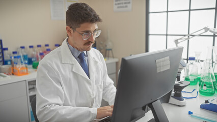 A young hispanic man in a lab coat working intently at a computer in a modern laboratory