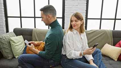 A woman sneakily glances at a man's smartphone as they sit back-to-back on a couch indoors.