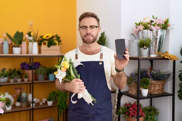 Middle age man with beard working at florist shop showing smartphone screen relaxed with serious expression on face. simple and natural looking at the camera.