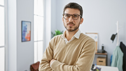 Confident young hispanic man with arms crossed standing in a modern office interior with a slight...