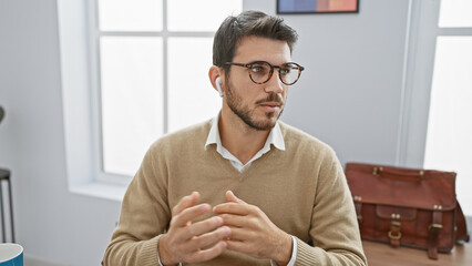 Handsome hispanic man with beard and glasses in office, wearing earbuds and sweater over shirt.
