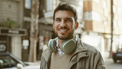 A handsome young hispanic man with a beard smiling on an urban street with headphones around his...