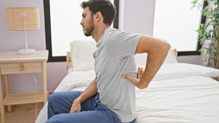 A young hispanic man with a beard sitting on a bed in a room, showing back pain.