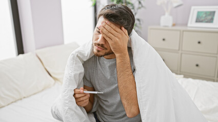 Hispanic man with beard looks unwell holding thermometer in bedroom setting.