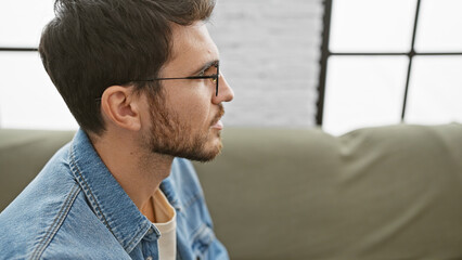 Profile of a thoughtful hispanic man in glasses wearing a denim shirt indoors, with a blurred...