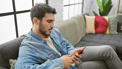 Handsome bearded hispanic man in casual denim attire using a smartphone while seated on a couch at...
