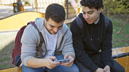Two young hispanic men share a moment looking at a smartphone while sitting together outdoors in a...