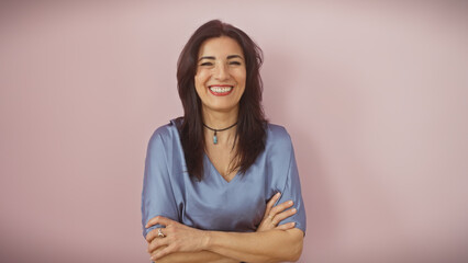 Smiling middle-aged hispanic woman with crossed arms standing against a pink background, exuding...