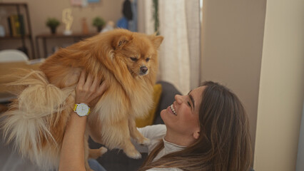 A smiling young hispanic woman holds her fluffy pomeranian indoors, depicting a cozy home scene.