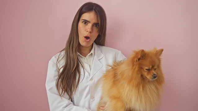Hispanic woman in white holding pomeranian against pink background shows surprise expression.