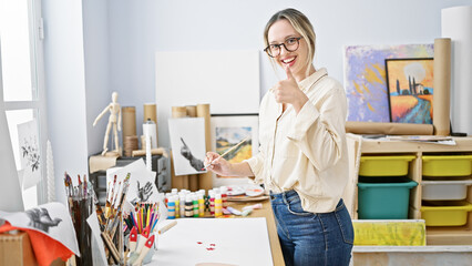 Young blonde woman artist drawing on paper doing thumb up gesture at art studio