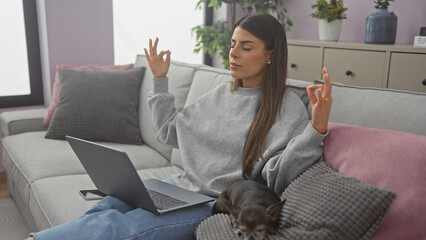 Young woman meditating on a couch with a laptop and a sleeping chihuahua in a cozy living room.