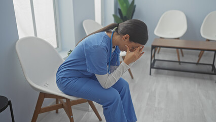 A distressed young woman in blue scrubs sits in a bright hospital room, covering her face with her...