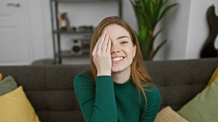 A young, smiling caucasian woman in a green sweater covers one eye playfully in a cozy, modern...