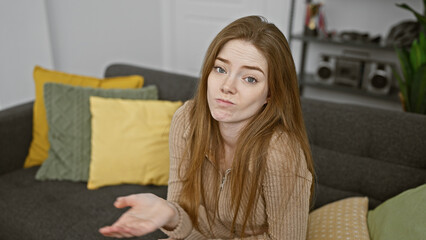 A confused young woman sits on a gray sofa indoors with colorful cushions, expressing doubt in a...