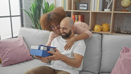 Interracial couple enjoys a cozy moment in a modern living room, sharing a surprise gift