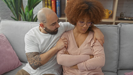 An interracial couple, a man and a woman, comforting each other on a grey sofa indoors