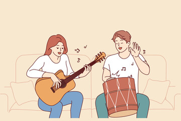 Cheerful friends man and woman playing musical instruments, sitting on sofa and using drum with guitar. Creative guy and girl enjoying playing musical hits during sunday relaxation