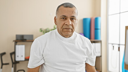 Mature hispanic man in a white shirt poses inside a rehabilitation clinic with medical equipment in...