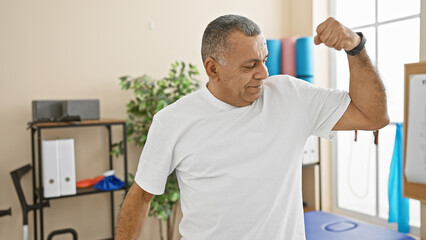 A mature hispanic man flexing muscles indoors at a rehabilitation clinic.