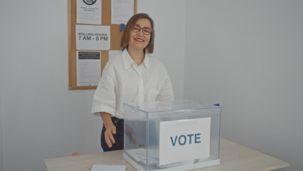 Smiling middle-aged woman supervising a transparent ballot box at an indoor electoral college room.