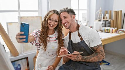 Obraz premium Smiling woman and tattooed man taking a selfie in an art studio, surrounded by paintings and supplies.