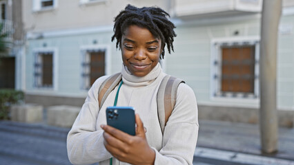 Smiling young black woman with dreadlocks using smartphone on city street