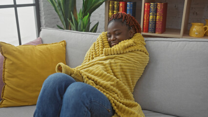 Obraz premium Young african american woman relaxing on a sofa wrapped in a yellow blanket in a cozy indoor setting