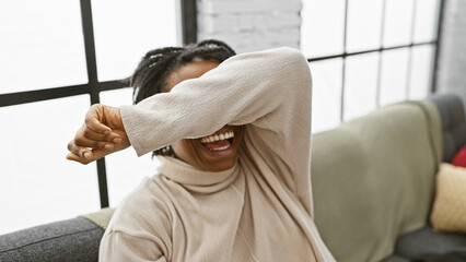 Laughing young black woman covering eyes with arm indoors, living room background, casual style,...