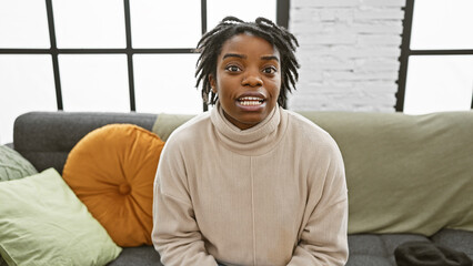 A surprised young woman with dreadlocks sits on a sofa in a modern living room, expressing emotion.