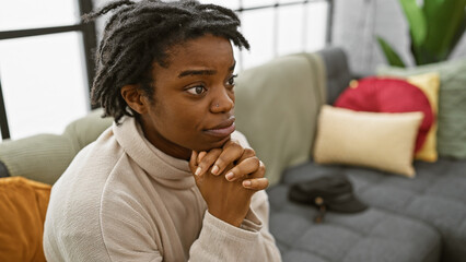 Pensive young adult african american woman with dreadlocks sitting thoughtfully on a sofa indoors.