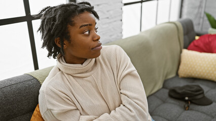 A thoughtful young woman with dreadlocks sits on a sofa indoors, providing a casual home setting.