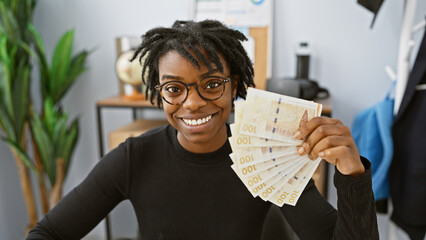Young woman in glasses holds danish currency in modern office setting, expressing cheerfulness.
