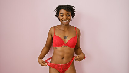 Smiling young black woman with dreadlocks, wearing red lingerie, stands against a pink isolated...