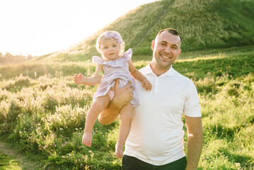 Father's Day. Happy baby hugging dad and standing on green grass in field in summer mountains. Dad...