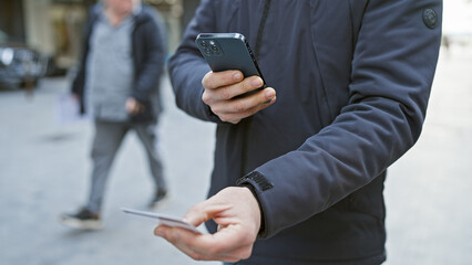 A young hispanic man captures a photo of a credit card on an urban street.