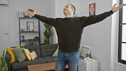Joyful hispanic man celebrating inside a modern apartment living room with arms raised
