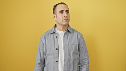 Hispanic man in grey shirt posing against an isolated yellow background looking away thoughtfully.