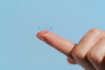 A finger of a female hand holds a blue transparent contact lens on a blue isolated background....