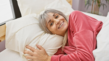 A smiling mature woman with grey hair wearing a pink top is lying comfortably in a bedroom setting...