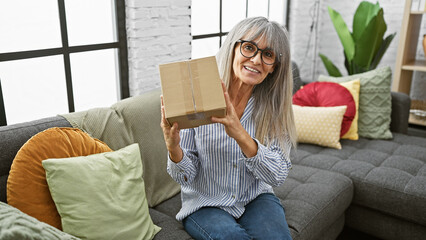 A smiling, grey-haired woman holding a package while seated on a sofa in a cozy living room...