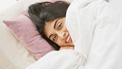 A serene south asian woman relaxing on a cozy bed with pillows in a bright bedroom setting.